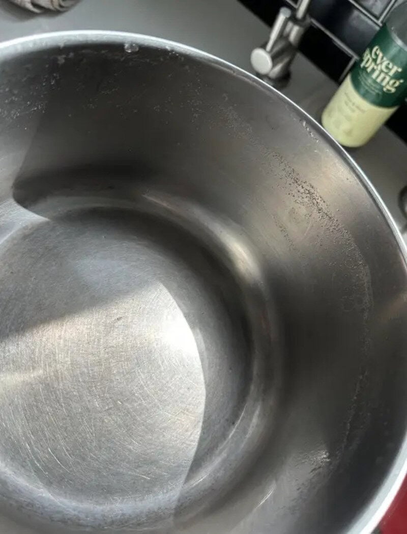 Empty stainless steel pot in a kitchen sink with soap and faucet in background.