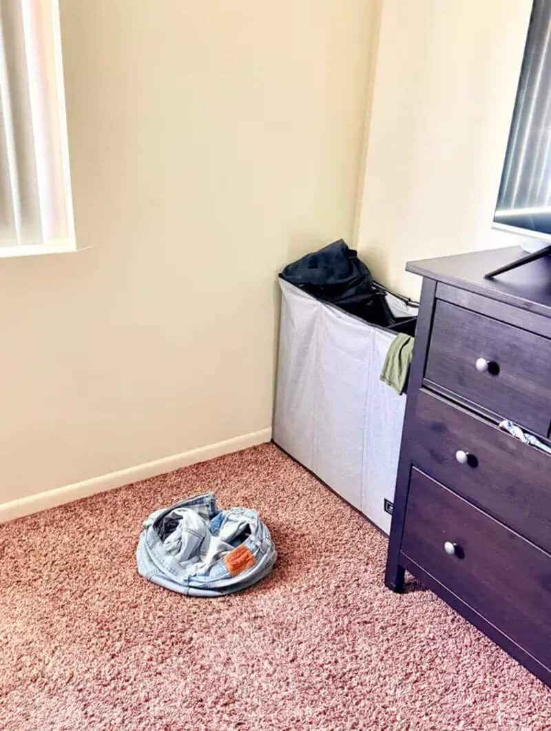Clothes hamper overflowing with laundry, on a pink carpet near a dresser.