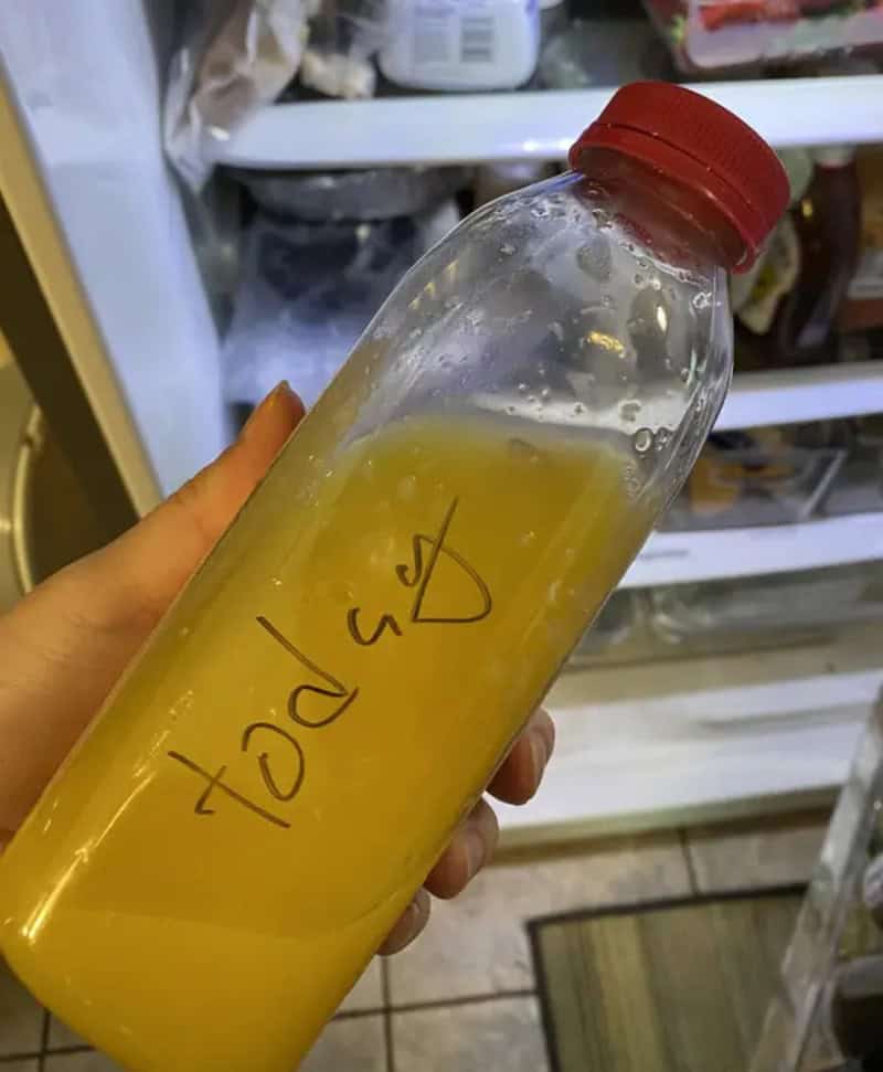 Bottle of yellow liquid with red cap in hand, fridge in background.