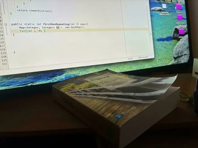 Computer screen displaying code and a book titled 'Java' on a desk.