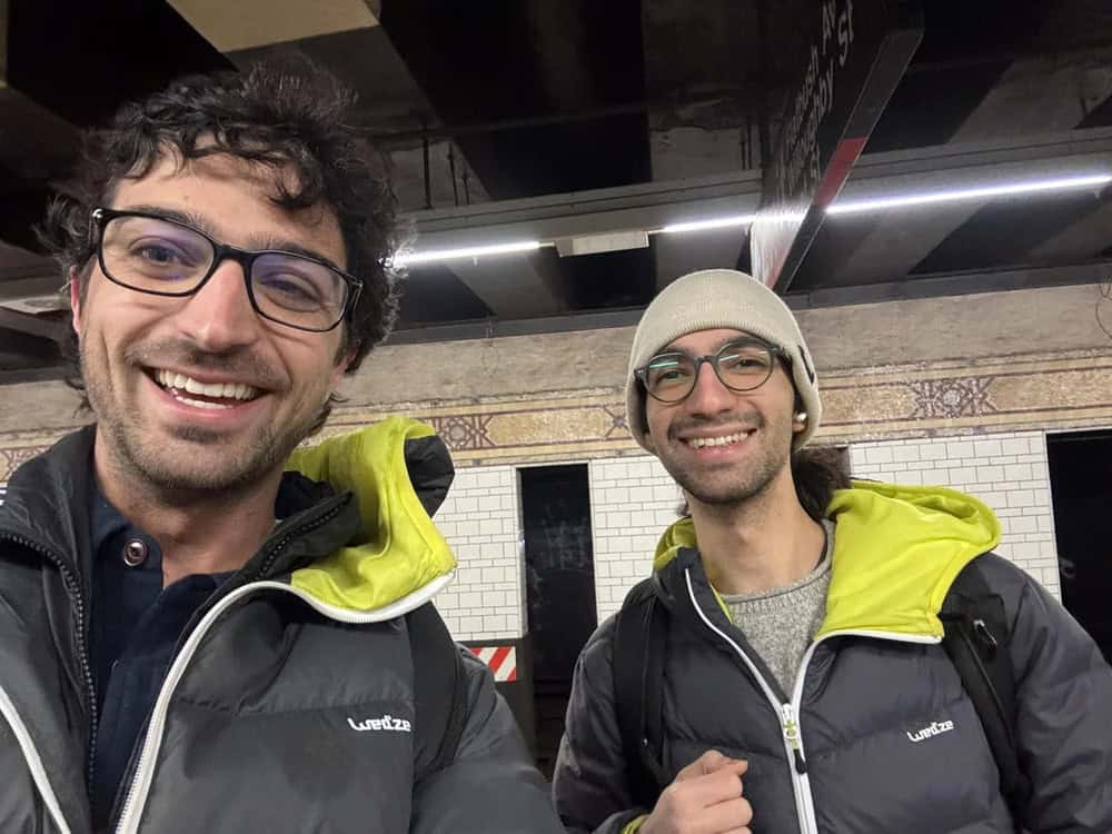 Two smiling men in jackets and glasses taking a selfie in a subway station.