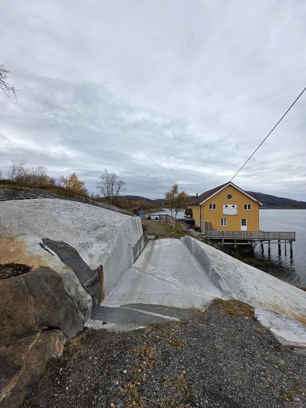 Concrete pathway leading to a yellow house on stilts over water under cloudy sky.
