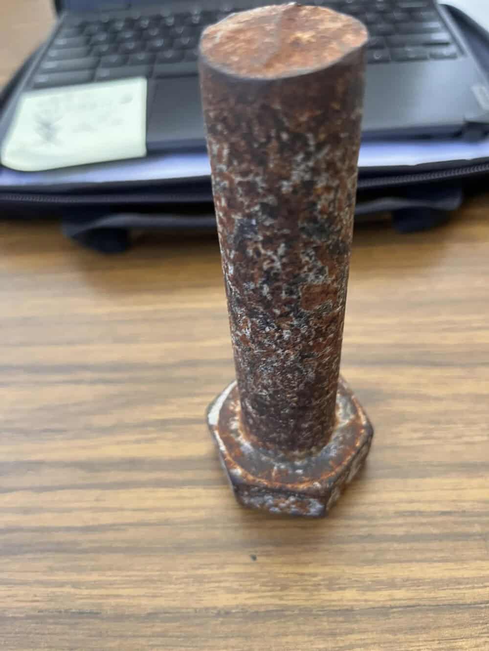 Rusty metal bolt standing upright on a wooden surface with a laptop in the background.