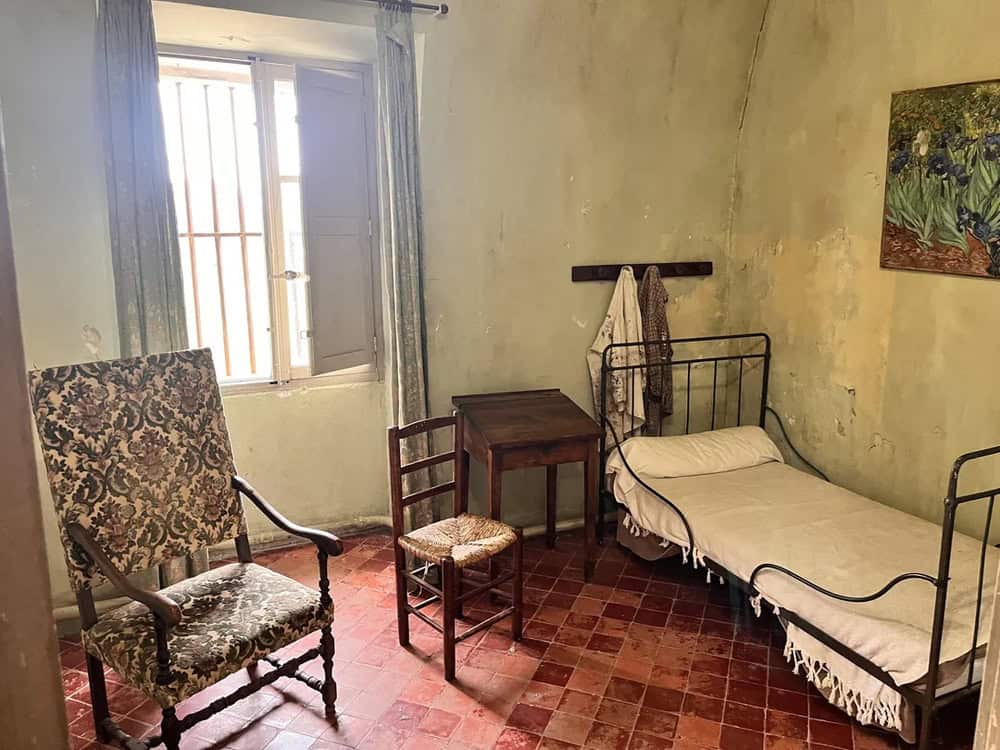 Old bedroom with a metal bed, wooden chair, armchair, and window with bars.