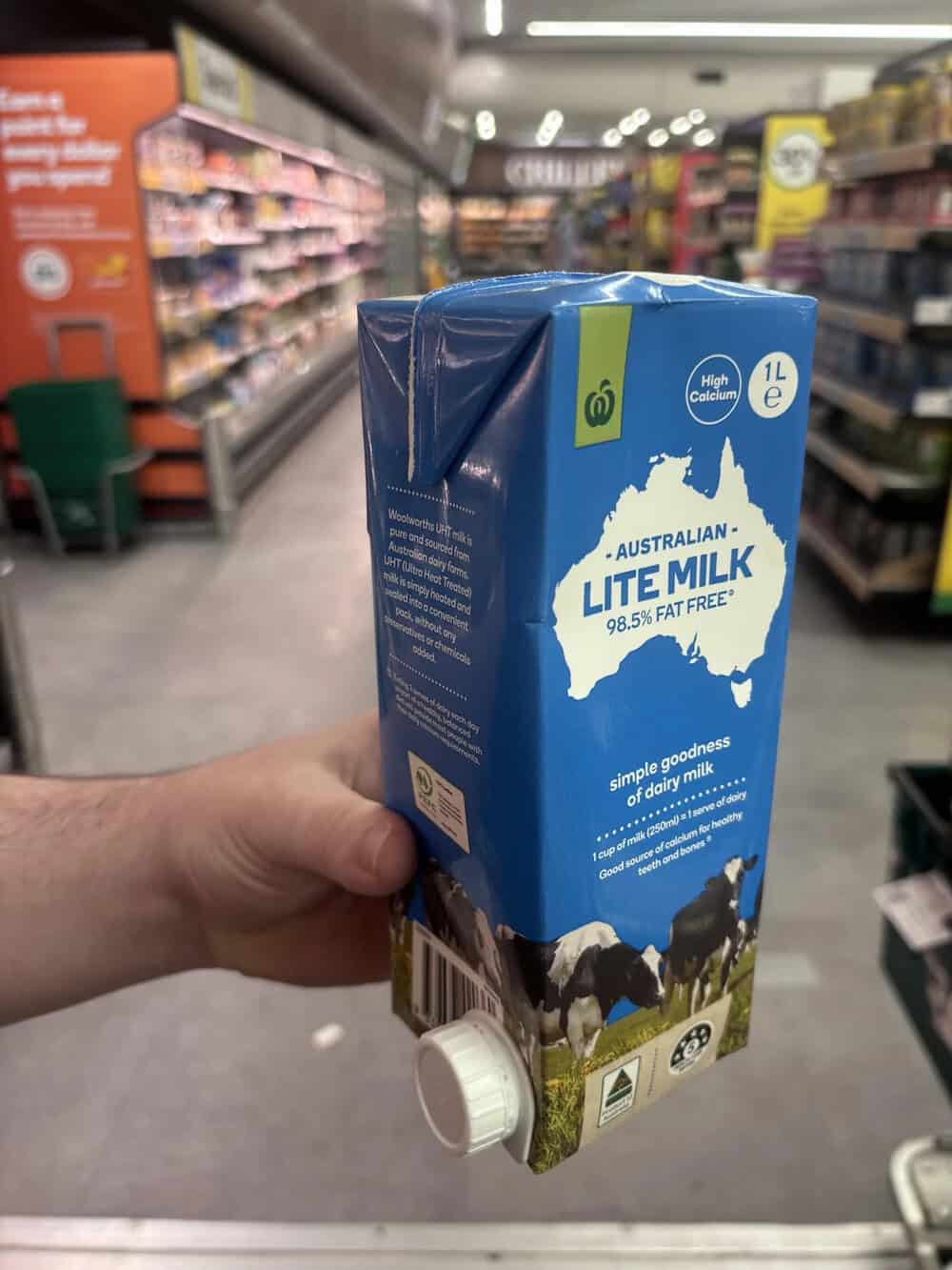 Hand holding a 1L carton of Australian Lite Milk in a grocery store aisle.