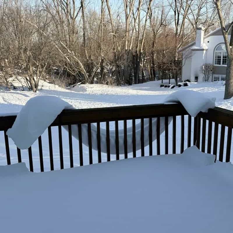 Snow-covered deck railing with trees and house in background during winter.