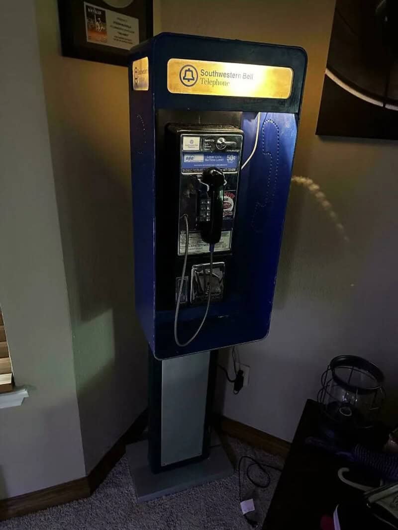 Public payphone booth with a blue exterior and yellow sign, located indoors.