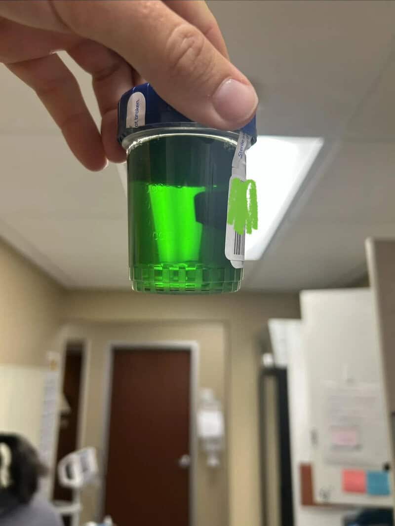 Hand holding a small container with bright green liquid in a room.