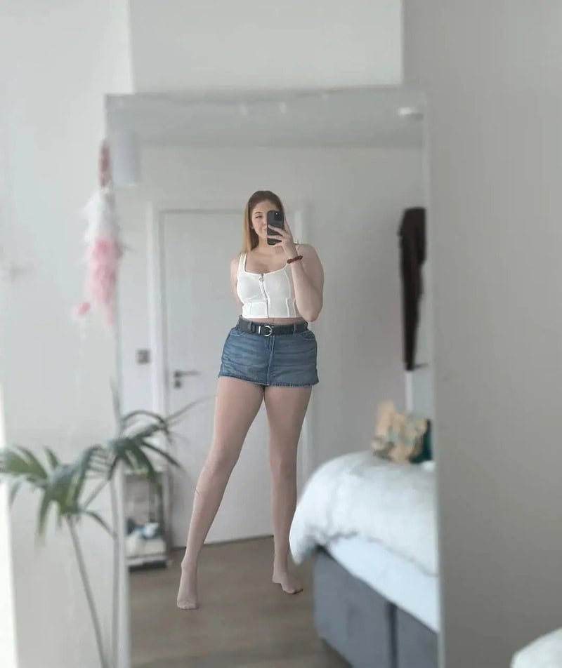 Young woman taking mirror selfie in a bedroom, wearing a white top and denim shorts.