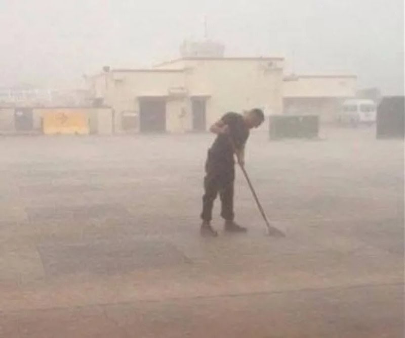 Person sweeping dust in a dusty, foggy environment with buildings in the background.
