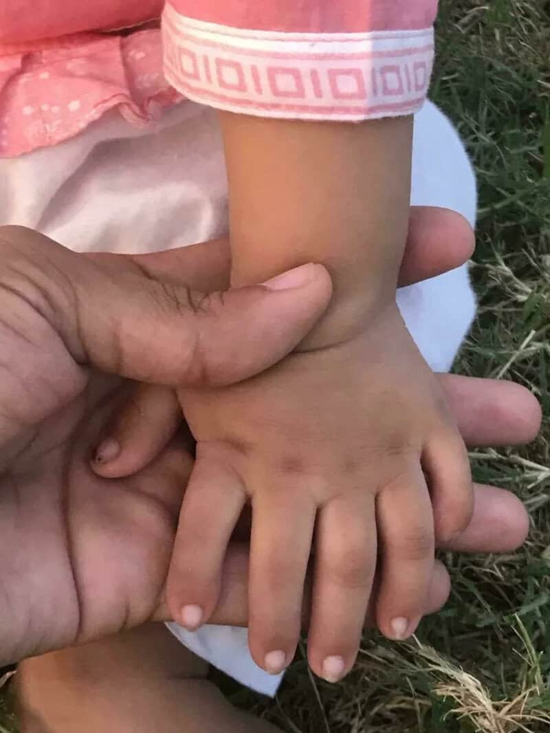 Child's hand holding an adult's finger, with grass in the background.