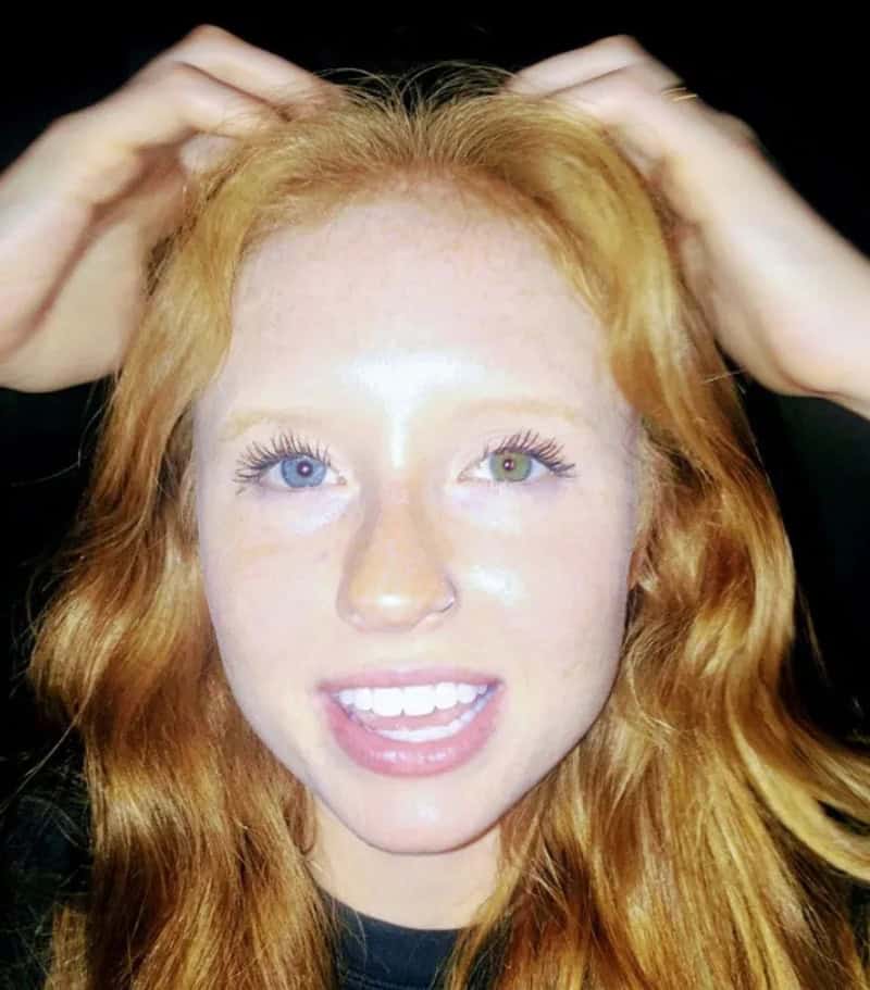 Woman with red hair, blue and green eyes, smiling, hands on her head.