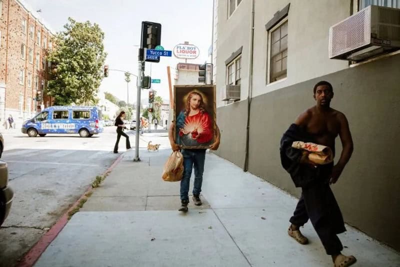 Person carrying a religious painting on a city sidewalk with a man holding a box.