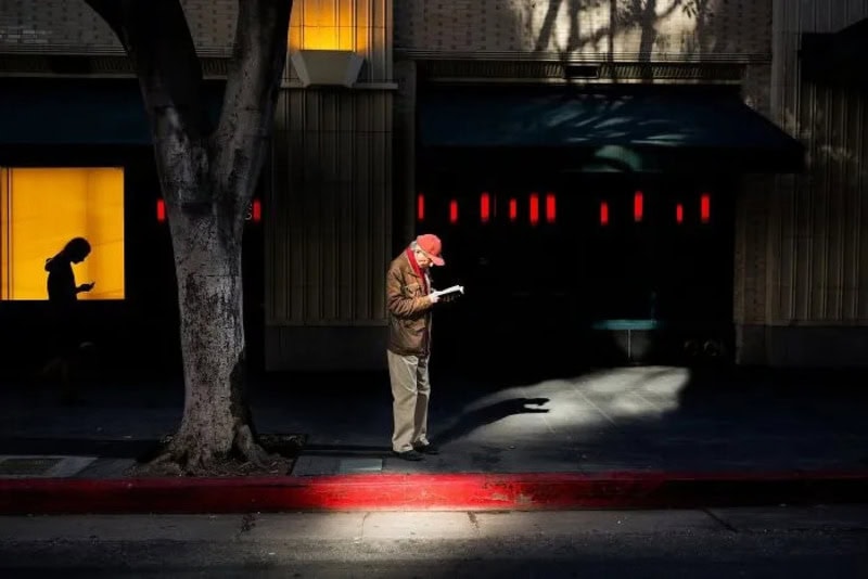 Man in coat reading a paper on a city sidewalk at night with a tree and building.