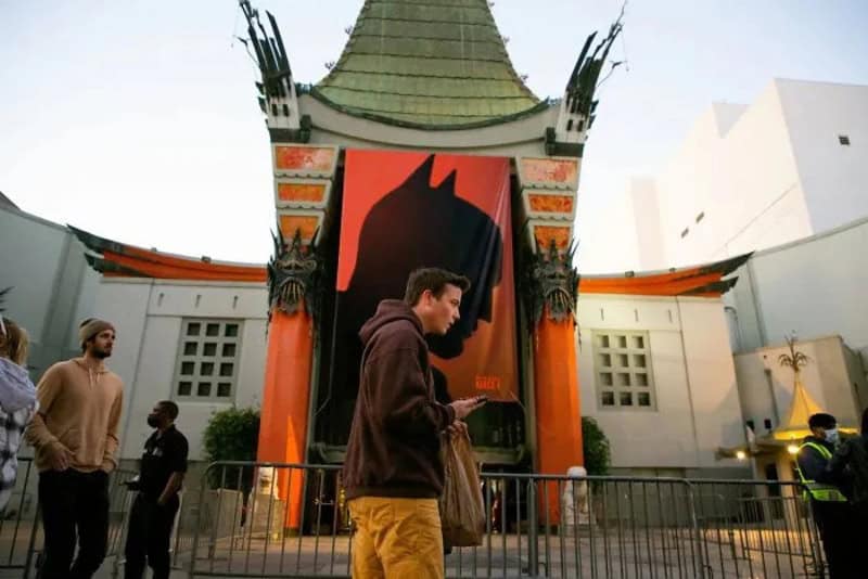 Man in brown hoodie and yellow pants using phone in front of Chinese theater.
