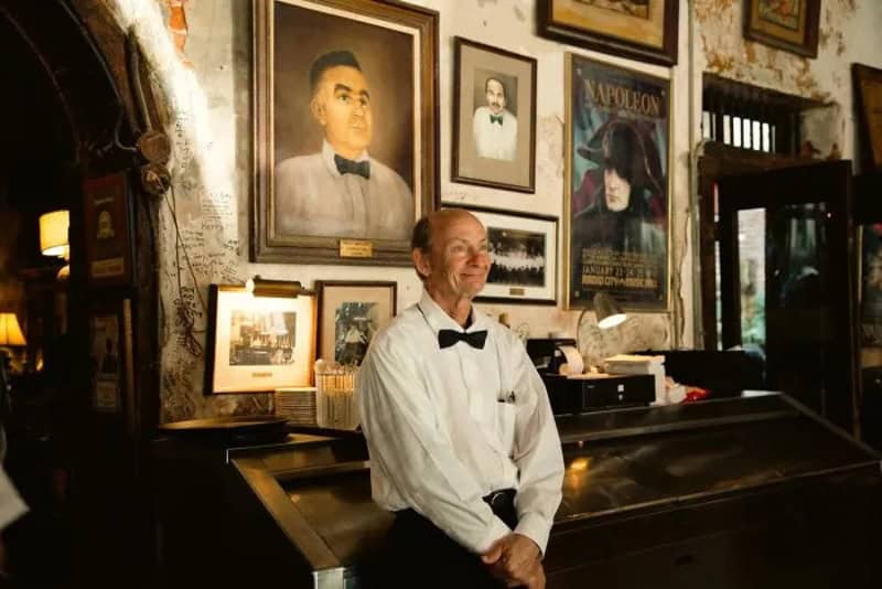 Man in white shirt and black bow tie standing in front of framed pictures and paintings.
