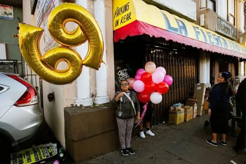 People with balloons on a city sidewalk near a store with a yellow awning.