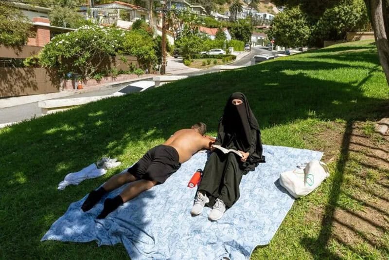 Person in niqab sitting on a blanket with a shirtless man lying beside her.