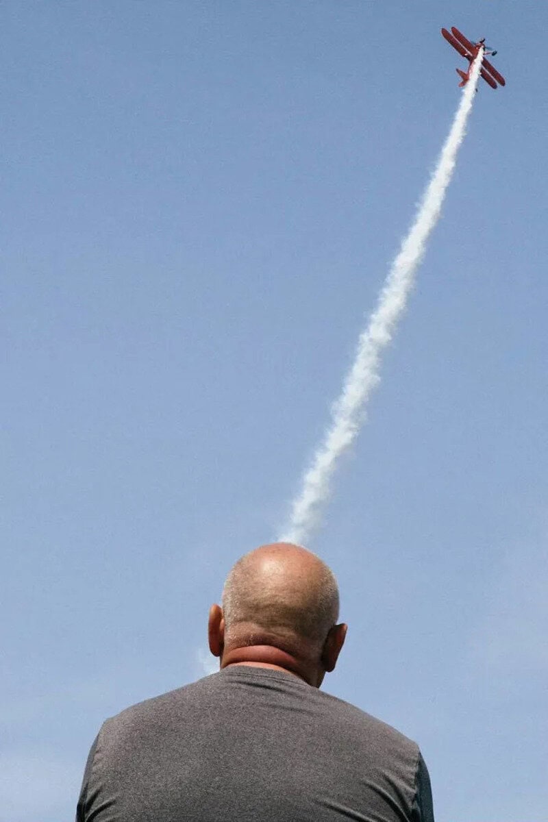 Bald man watching a red airplane flying high with a trail of white smoke.