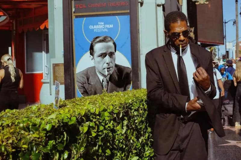 Man in black suit and sunglasses on street with poster of a man in a suit.