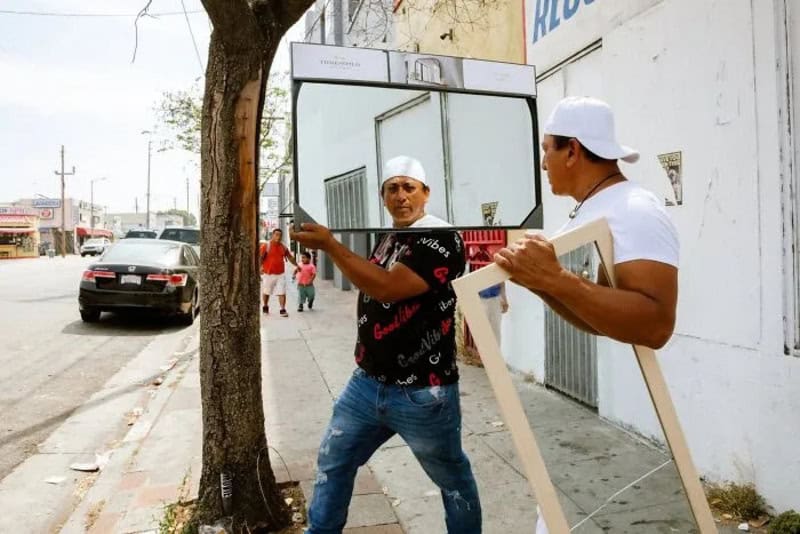 Two men in white caps look at a mirror on a sidewalk in Los Angeles.