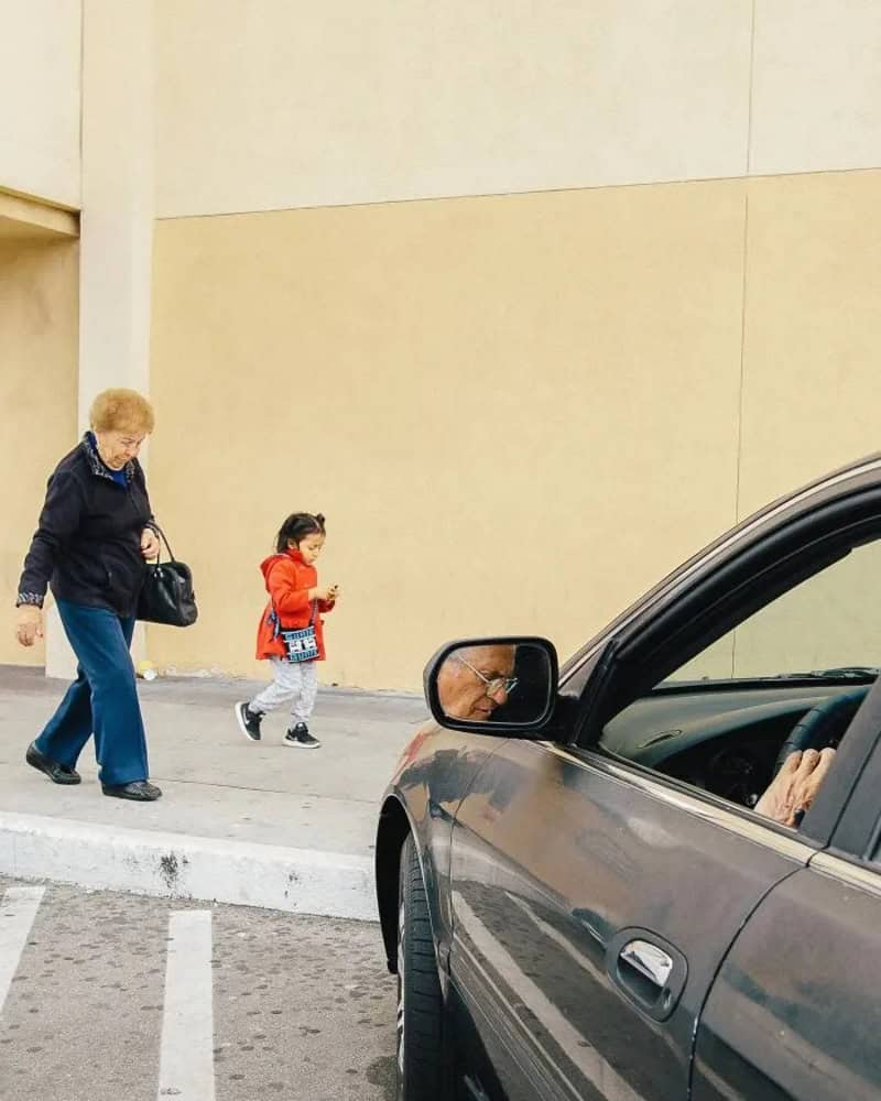 An elderly woman and a young girl walk past a parked car on a city street.