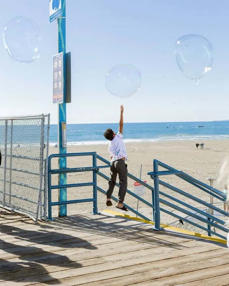 Boy reaching for balloons at the beach with ocean in background.