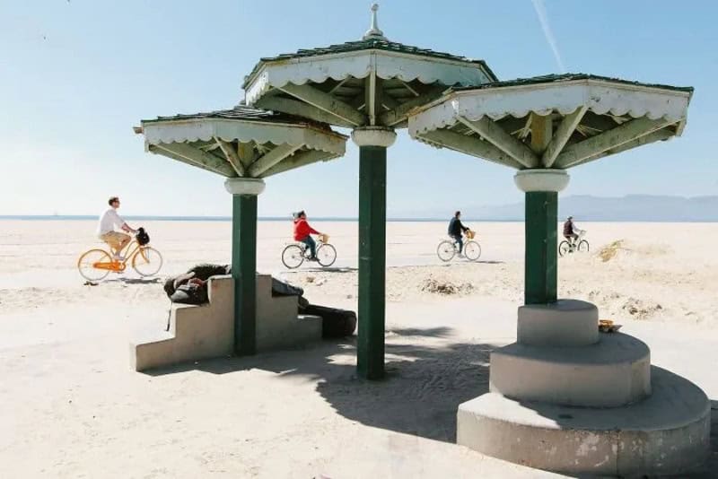 People riding bikes under beach umbrellas on a sandy shore.