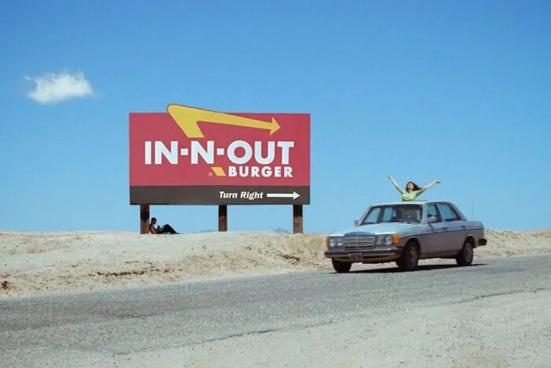 Person with arms raised in front of In-N-Out Burger sign on roadside.