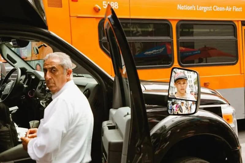 Man in white shirt sitting in a vehicle with a bus in the background.