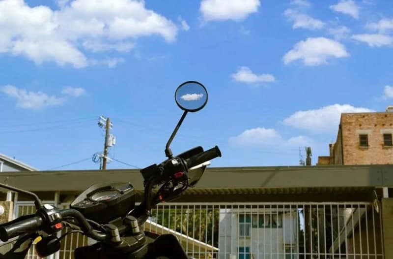 Motorcycle handlebar with a round mirror reflecting the sky and clouds.