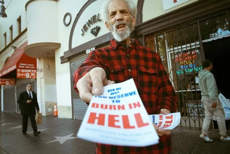 Elderly man in red plaid shirt holding a flyer on the street.