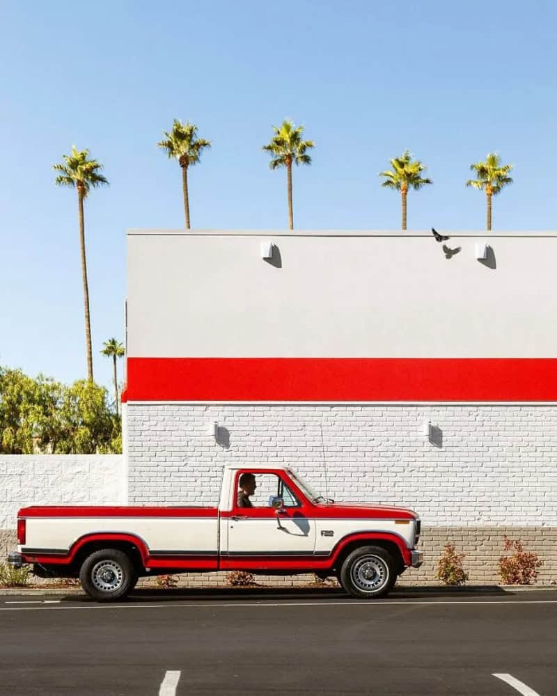 Red and white pickup truck parked in front of a white wall with palm trees in the background.