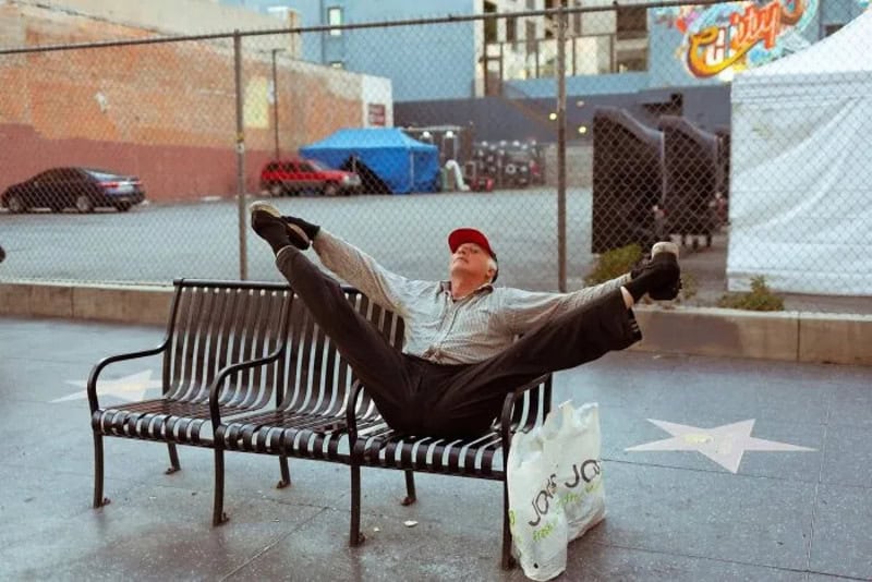 Man stretching on a park bench in Los Angeles with a shopping bag nearby.