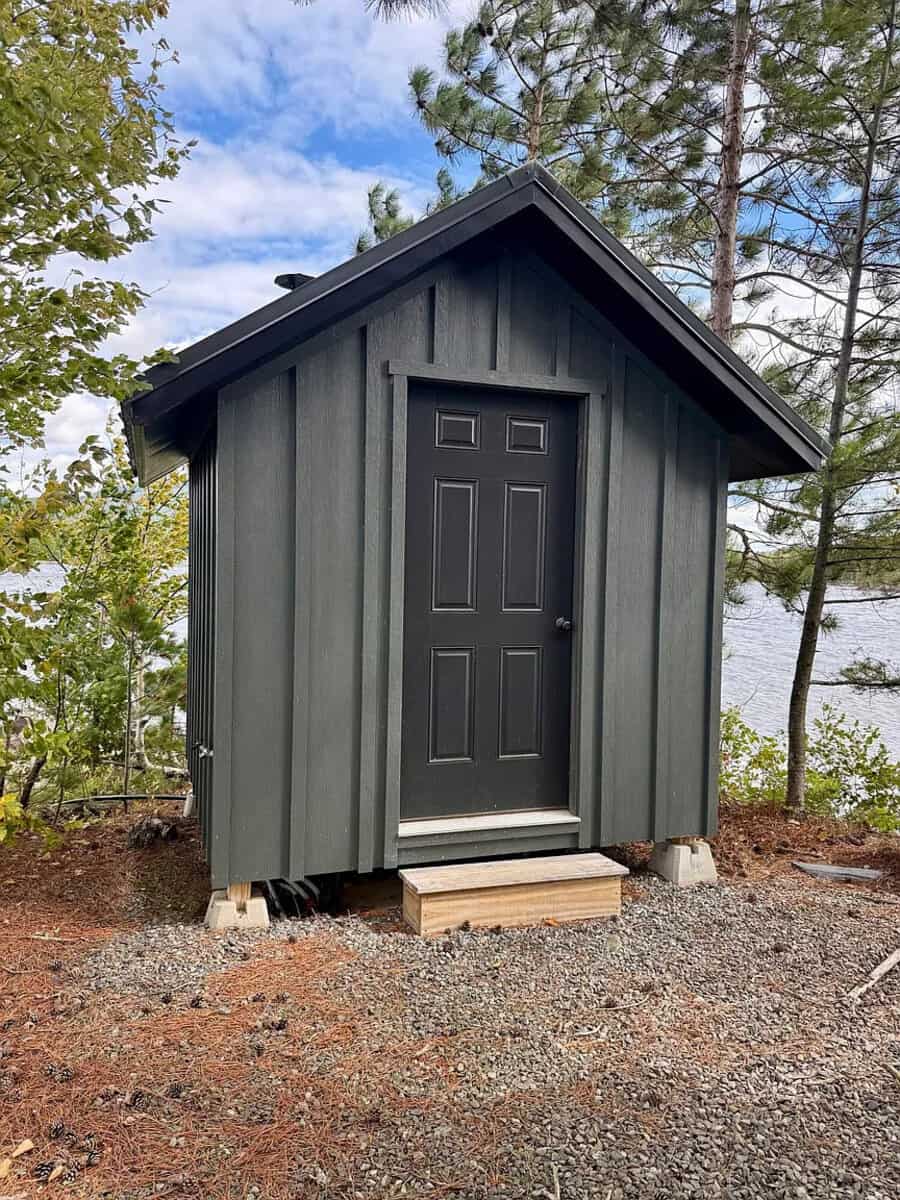 Small dark gray shed with a door, surrounded by trees and lake in background.