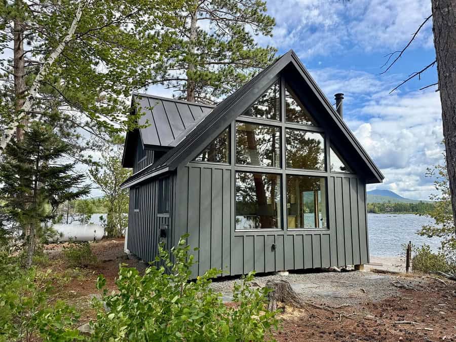 Modern black house with large windows by a lake, surrounded by trees.