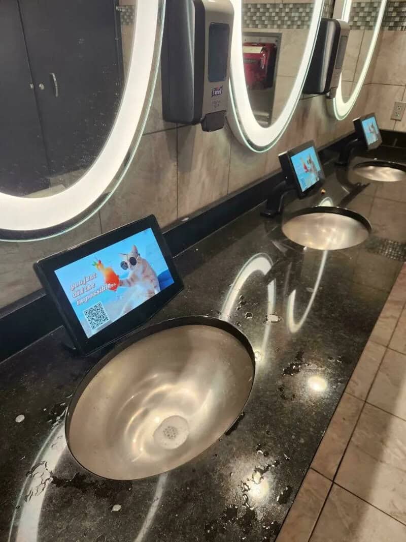 Public restroom sinks with digital screens and mirrors above, wet countertop.