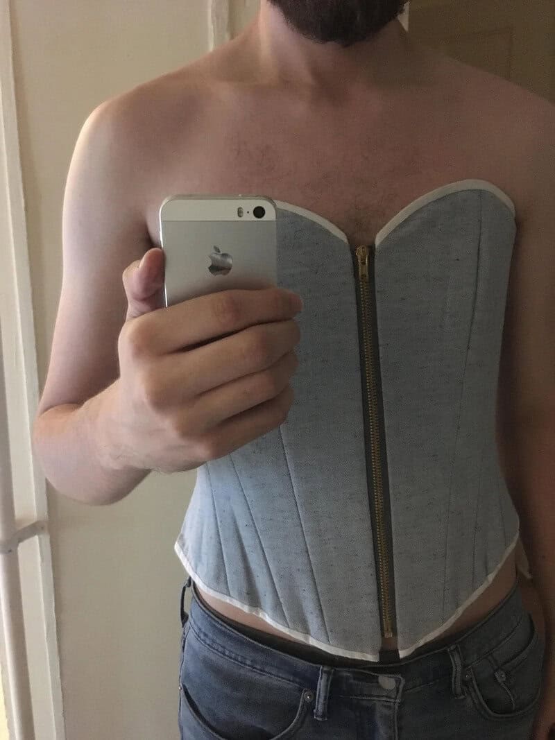 Person taking a mirror selfie wearing a gray corset with a zipper in the front.