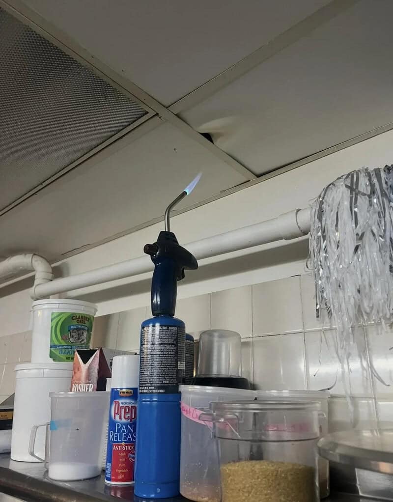 A small kitchen torch with a blue handle and a lit flame, on a kitchen counter.