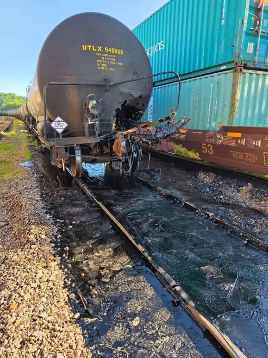 Damaged black tanker railcar on tracks with burnt and melted parts.