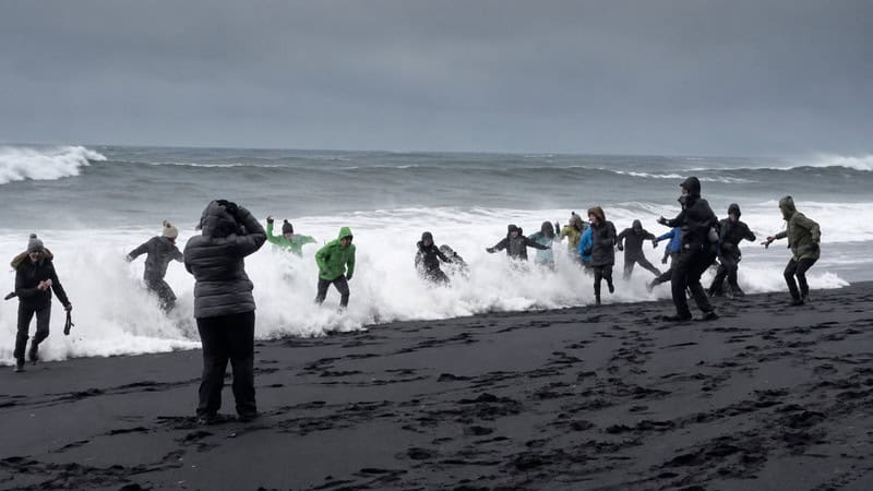 People on a dark beach playing in the ocean waves under a cloudy sky.