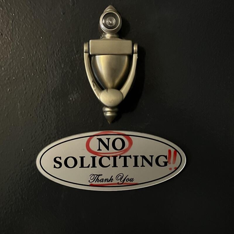 No soliciting sign on a dark wall with a doorbell above it.