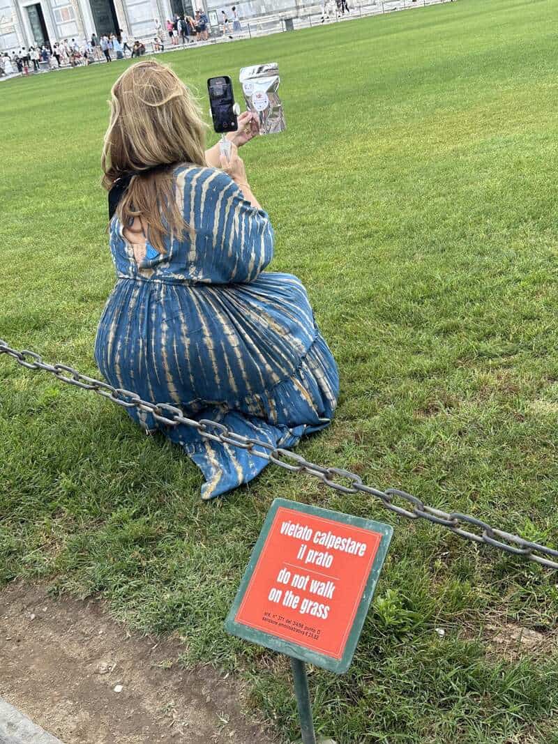 Woman in blue dress taking a photo on her phone on grass, with a sign in front that says do not walk.