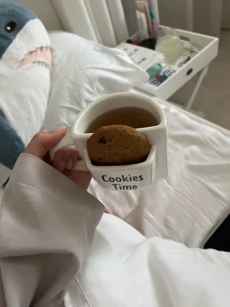 Person holding a mug with cookies and tea, on a bed with a white blanket.