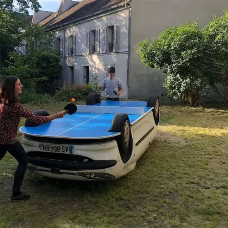 People playing table tennis on a flipped car in a yard.
