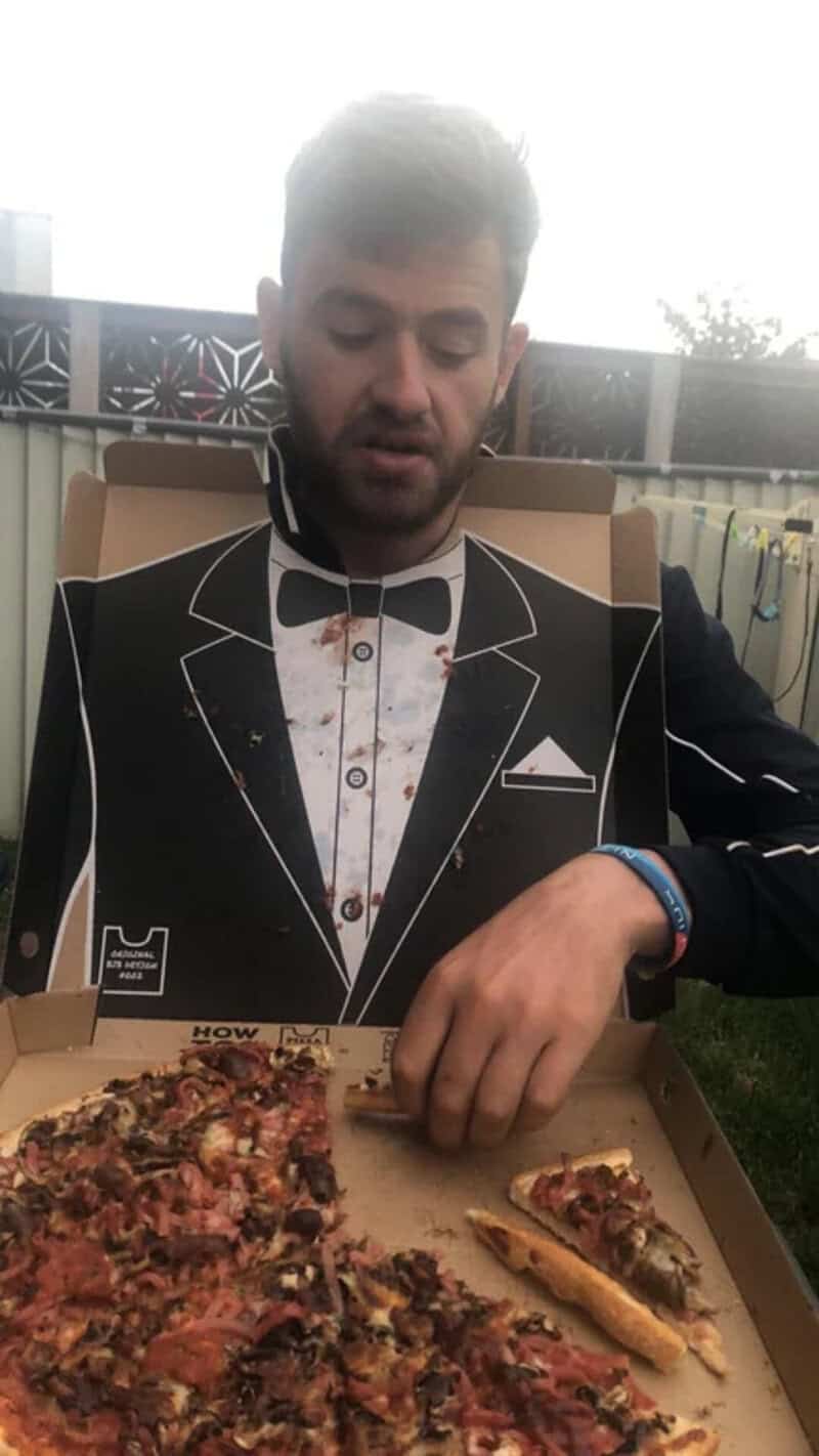 Man in tuxedo costume with pizza box, outdoor setting, overcast sky.