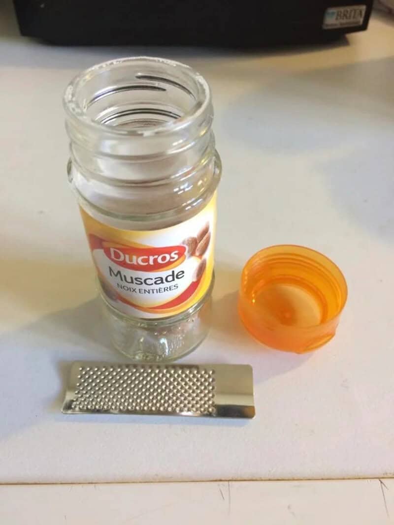 A small grater, a bottle of Ducros Muscade, and an orange cap on a white surface.
