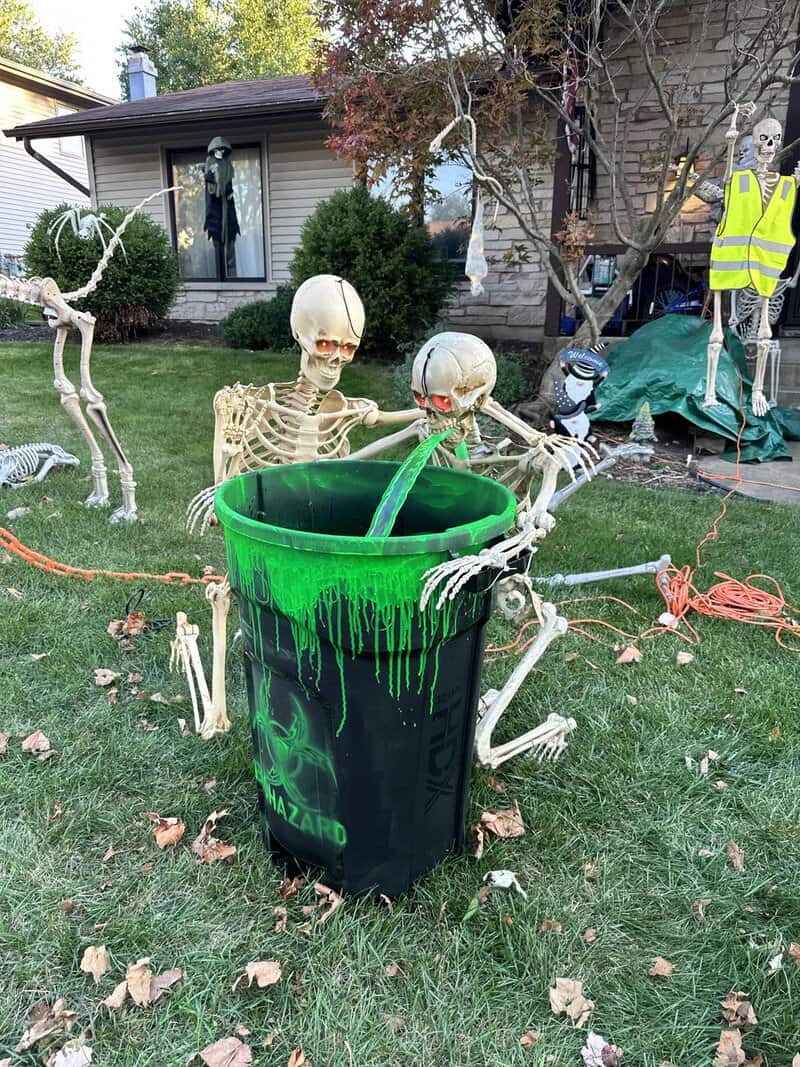 Two skeletons leaning over a green trash can in a Halloween yard display, with other skeletons and a.