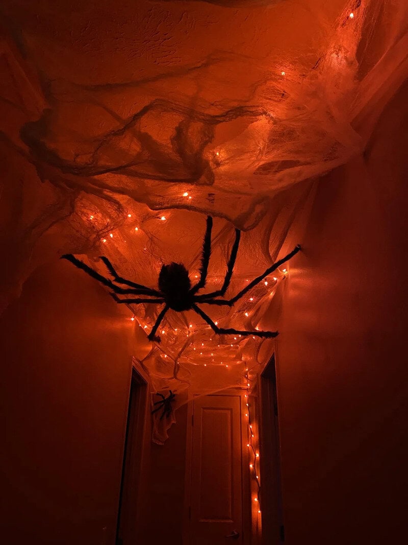 Large black spider decoration with orange Halloween lights along a hallway ceiling, cobwebs overhead.