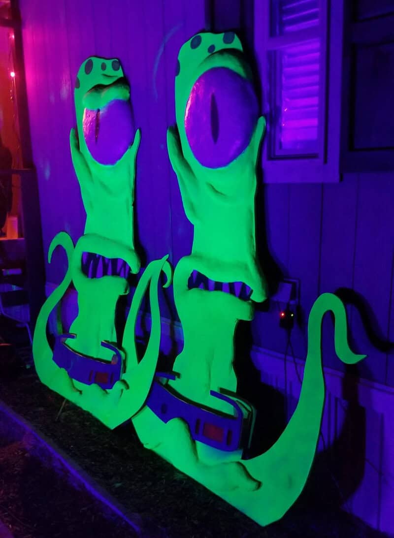 Two neon-green inflatable aliens with large purple eyes mounted on a house, under purple lighting.